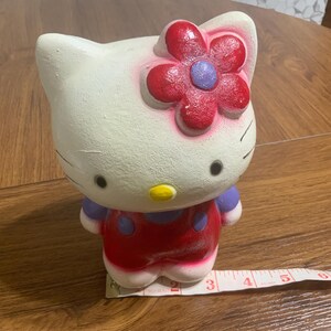 May include: A ceramic Hello Kitty figurine, painted in white, red, and purple. The figurine has a red flower on its head and is approximately 3 inches tall. The figure is standing on a wooden surface.