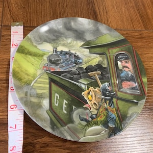 May include: A decorative plate featuring a whimsical train scene. The plate depicts a green train with a conductor and a cartoonish creature, with another train in the background. The plate is approximately 8 inches in diameter.