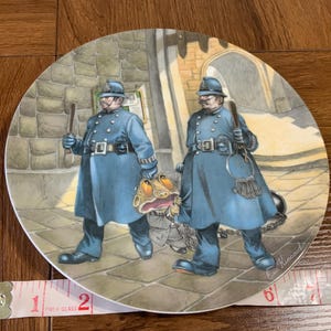 May include: A decorative plate featuring an illustration of two police officers in blue uniforms, holding batons, and walking with a frog. The plate has a glossy finish and a detailed, colorful design.