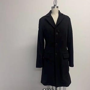 May include: A black overcoat displayed on a mannequin. The coat has a notched collar, three buttons, and two front pockets. The coat is knee-length and has long sleeves. The mannequin is on a metal stand against a white background.