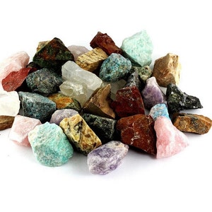 May include: A collection of various raw gemstones in shades of red, pink, green, blue, brown, and purple. The stones are of different sizes and shapes, with a variety of textures and patterns. The gemstones are scattered on a white background.