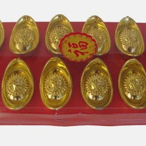 May include: A display of twelve gold-coloured ingots presented on a red rectangular base. Each ingot features an intricate design. A circular red and gold emblem with Chinese characters is positioned centrally among the ingots.