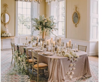 Puede incluir: Una mesa larga y rectangular con un mantel de lentejuelas rosa claro está preparada para una cena formal. La mesa está decorada con flores blancas, velas y detalles dorados. Las sillas son doradas y tienen un diseño sencillo.
