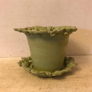 May include: A light green cast iron planter with a decorative, scalloped rim and a matching saucer. The planter has a textured, striped pattern.
