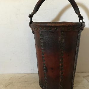 May include: A vintage leather bucket with a curved handle. The bucket is a dark brown color, with metal rivets along the seams. The handle is also made of leather.