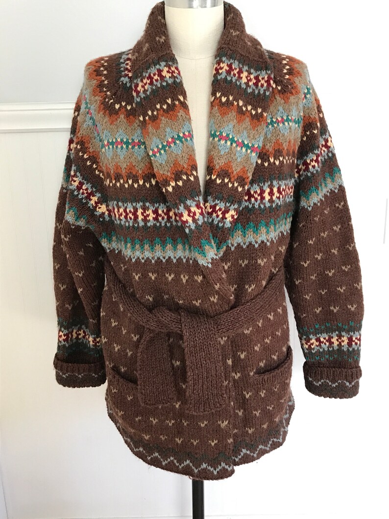 Puede incluir: Un c&aacute;rdigan de punto marr&oacute;n con un estampado geom&eacute;trico colorido y un cintur&oacute;n de anudar. El c&aacute;rdigan tiene un cuello chal y bolsillos.