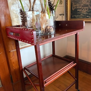 May include: A vintage wooden bar cart with a red leather handle and brass studs. The cart has two shelves and sits on wheels. Glass vases with antlers, flowers, and pinecones are displayed on the top shelf. A framed painting hangs in the background.