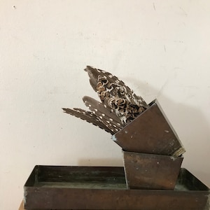 May include: A set of three brown metal trays with a bird feather arrangement inside. The trays are stacked on top of each other and the feather arrangement is in the top tray.