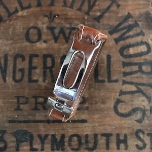 May include: A silver and brown leather watch band with a buckle closure. The band is on a wooden surface with the text "O.W. Paint Works" printed on it.