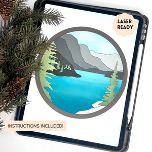 May include: A digital illustration of a mountain lake scene with a blue lake, gray mountains, and green trees. The image is in a circle and is labeled "Laser Ready" and "Instructions Included!"