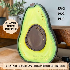 May include: A five-layer digital cut file of an avocado with a brown pit. The avocado is green with a yellow outline. The text "Kendra's Got Wood" is visible at the bottom of the image. The text "SVG, PNG, PDF, 5 LAYER DIGITAL CUT FILE, CUT ON LASER OR SCROLL SAW - INSTRUCTIONS FOR BOTH INCLUDED!" is also visible in the image.