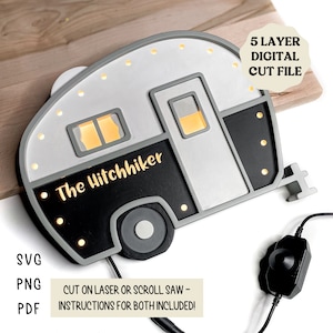 May include: A black and white wooden cutout of a camper trailer with the text "The Hitchhiker" on the side. The camper has a light gray body and black trim. The cutout is lit up with small white lights. The image includes the text "5 Layer Digital Cut File" and "Cut on laser or scroll saw - Instructions for both included!"