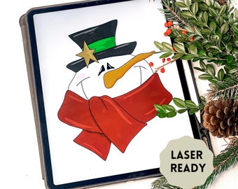 May include: A digital illustration of a snowman wearing a green and black hat with a gold star on top and a red scarf. The image is on a tablet screen with the text "LASER READY" on a beige circle.