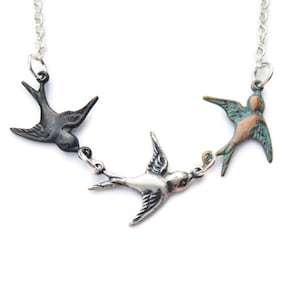 May include: A silver chain necklace with three bird charms. The birds are in flight and are made of metal with different finishes. One bird is black, one is silver, and one is a copper colour with a turquoise patina.