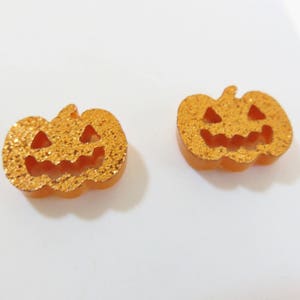 Puede incluir: Dos pendientes de Halloween de color naranja en forma de calabaza con detalles de purpurina y una cara de calabaza.