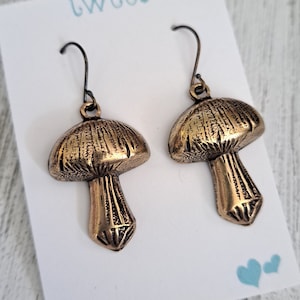 Mushroom earrings in antiqued brass with antiqued brass ear wires. Shown on a white earring card that says Twee and has 2 little hearts in the lower right corner. Text & hearts are aqua colored. Earring card is resting on white wood background.