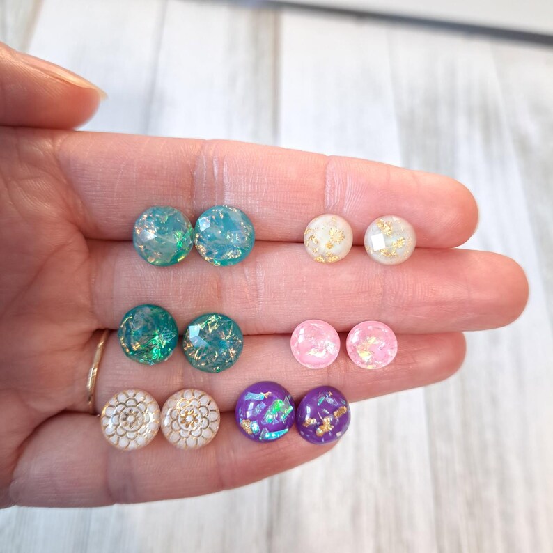 May include: A collection of round stud earrings in various colors, including teal, green, pink, white, and purple. Some earrings feature gold leaf accents and decorative patterns. The earrings are displayed on a hand.