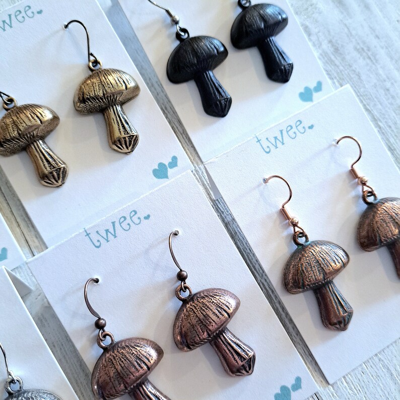 Close up of several pairs of mushroom earrings on white and aqua earring cards. Detail is shown on antiqued brass, matte black, antiqued copper, and copper verdigris versions.