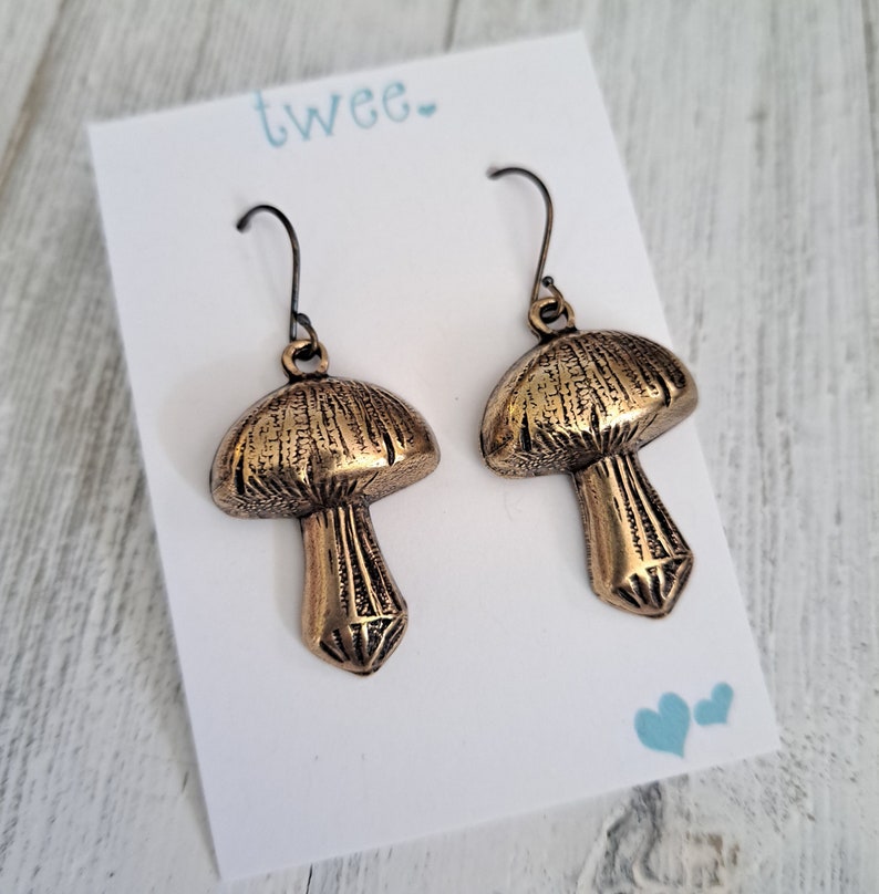 Mushroom earrings in antiqued brass with antiqued brass ear wires. Shown on a white earring card that says Twee and has 2 little hearts in the lower right corner. Text & hearts are aqua colored. Earring card is resting on white wood background.