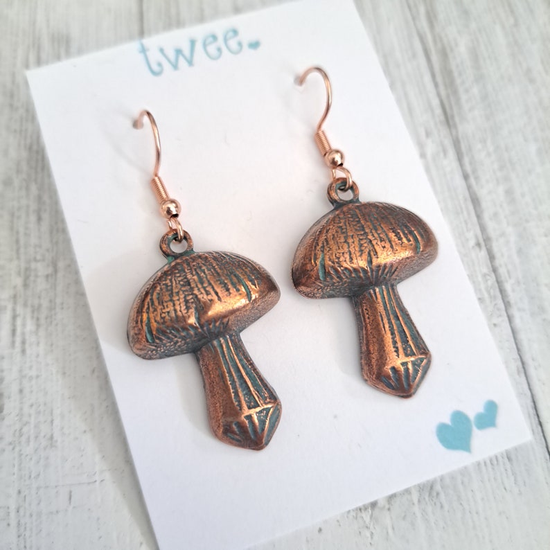 Mushroom earrings in copper with a verdigris patina and bright copper ear wires. Shown on a white earring card that says Twee and has 2 little hearts in the lower right corner. Text & hearts are aqua colored. Earring card is on white wood background.