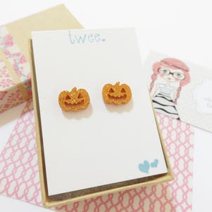 Puede incluir: Un par de pendientes de calabaza naranja con purpurina en una peque&ntilde;a caja blanca con la palabra "twee" impresa en la parte delantera.