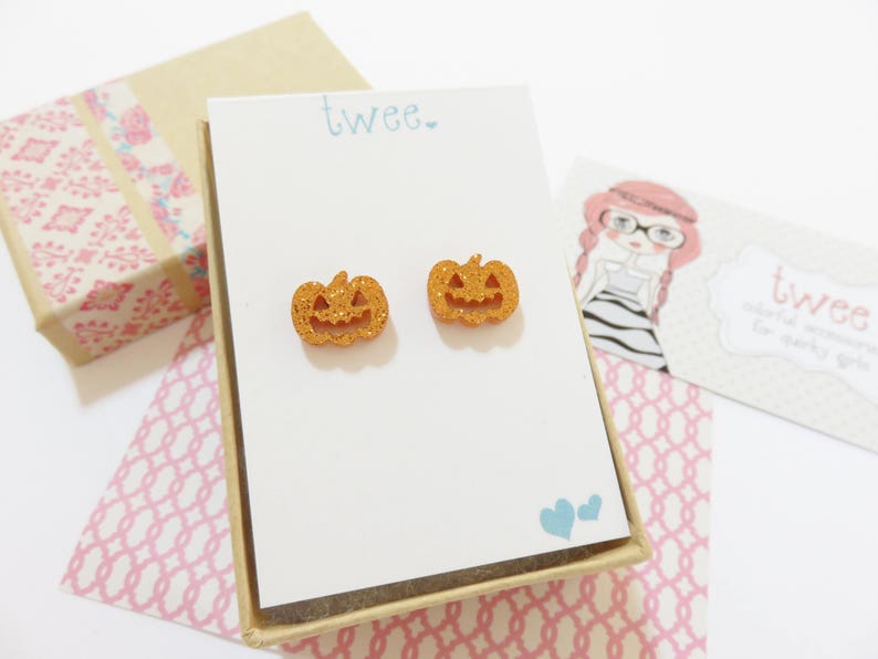 Puede incluir: Un par de pendientes de calabaza naranja con purpurina en una peque&ntilde;a caja blanca con la palabra "twee" impresa en la parte delantera.