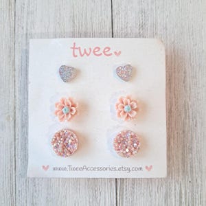 May include: A set of six stud earrings on a white card. The earrings include two silver glitter hearts, two peach flower studs with blue centers, and two round, pink glitter studs. The card has the word "twee" in pink and the Etsy shop URL.