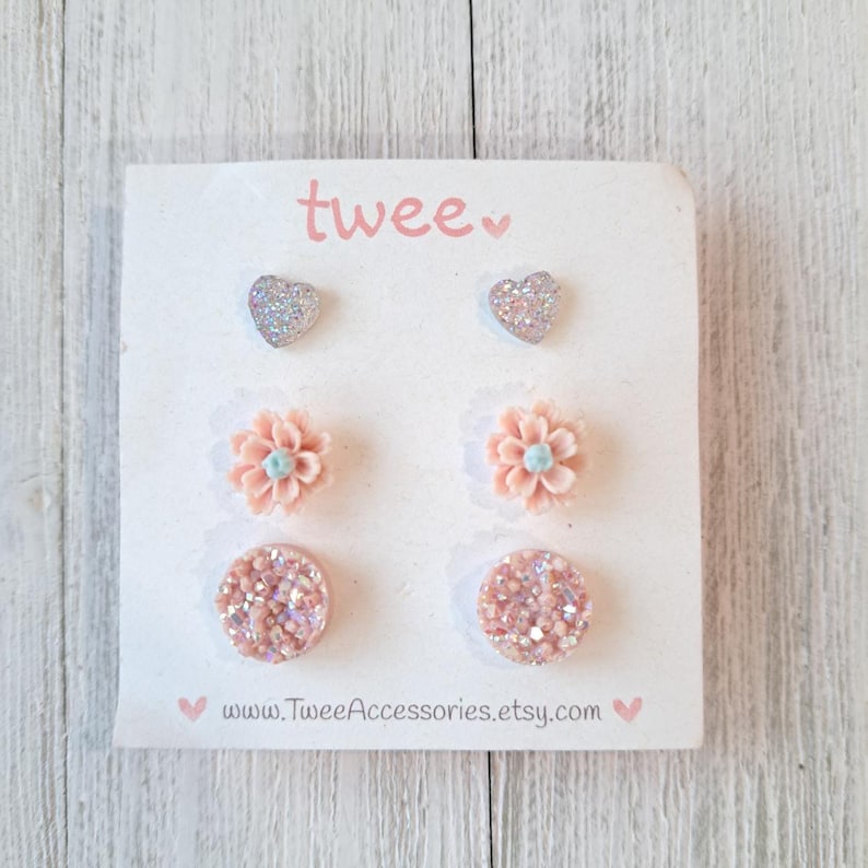 May include: A set of six stud earrings on a white card. The earrings include two silver glitter hearts, two peach flower studs with blue centers, and two round, pink glitter studs. The card has the word "twee" in pink and the Etsy shop URL.