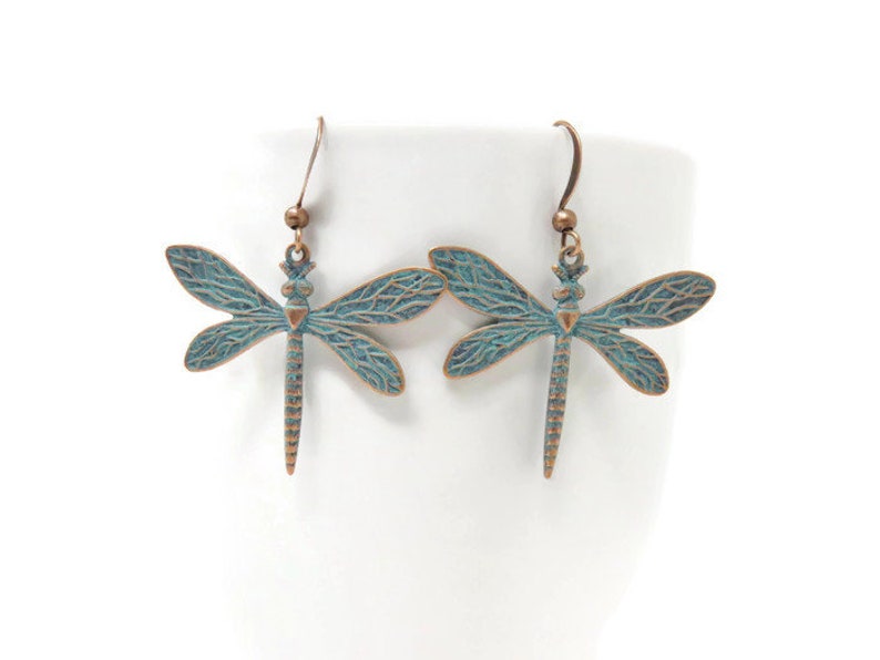 May include: A pair of copper-toned dragonfly earrings with a teal blue patina finish. The earrings have a delicate, detailed design and are perfect for adding a touch of nature to any outfit.