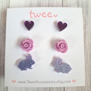 May include: A set of six stud earrings on a white card. The earrings include two purple heart-shaped studs, two light purple rose studs, and two glittery purple bunny studs. The card has the word "twee" in pink and the Etsy shop's URL.