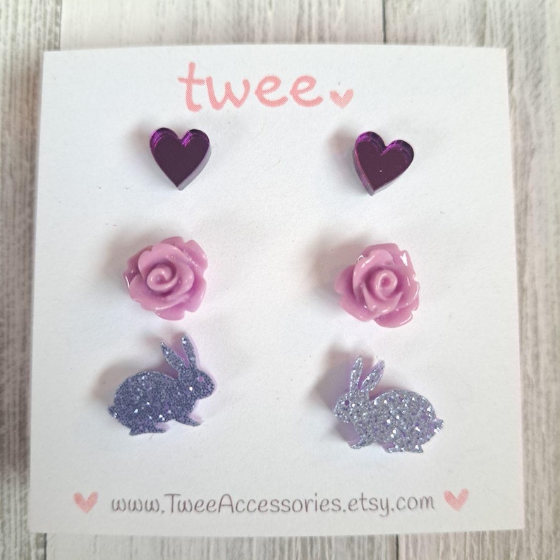 May include: A set of six stud earrings on a white card. The earrings include two purple heart-shaped studs, two light purple rose studs, and two glittery purple bunny studs. The card has the word "twee" in pink and the Etsy shop's URL.