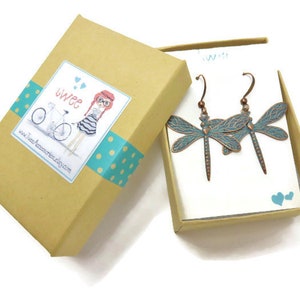 May include: A pair of dragonfly earrings in a gift box. The earrings are made of copper and have a turquoise blue enamel finish. The box is brown with a white label that says "twee" and has a cartoon illustration of a girl riding a bicycle.