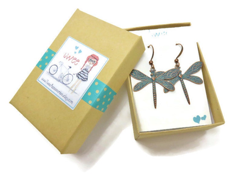 May include: A pair of dragonfly earrings in a gift box. The earrings are made of copper and have a turquoise blue enamel finish. The box is brown with a white label that says "twee" and has a cartoon illustration of a girl riding a bicycle.