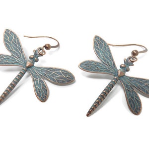 May include: A pair of dangle earrings featuring a dragonfly design. The dragonflies are made of metal with a copper finish and have a blue-green patina. The earrings have a hook closure.