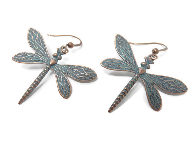 May include: A pair of dangle earrings featuring a dragonfly design. The dragonflies are made of metal with a copper finish and have a blue-green patina. The earrings have a hook closure.