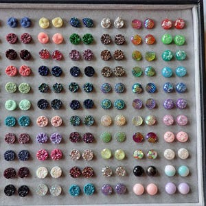 May include: A display of colorful stud earrings in various colors, including blue, pink, green, and gold. The earrings have a textured, glittery appearance and are arranged in a grid pattern on a gray surface.
