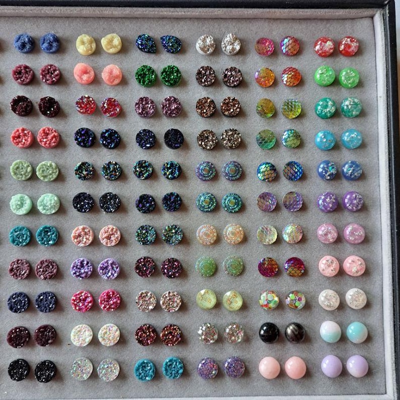 May include: A display of colorful stud earrings in various colors, including blue, pink, green, and gold. The earrings have a textured, glittery appearance and are arranged in a grid pattern on a gray surface.
