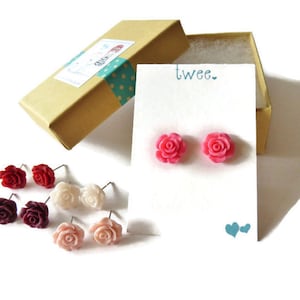 May include: A collection of rose-shaped stud earrings in various colors, including red, pink, and cream, displayed on a white card with the word "twee." and a pair of blue hearts. The earrings are presented with a small, open, tan gift box.