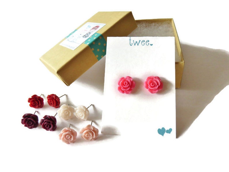 May include: A collection of rose-shaped stud earrings in various colors, including red, pink, and cream, displayed on a white card with the word "twee." and a pair of blue hearts. The earrings are presented with a small, open, tan gift box.