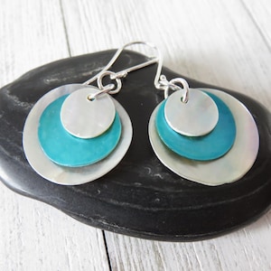 May include: Silver-tone earrings with a layered design. Each earring features three circular elements: a small silver disc, a medium teal disc, and a large silver disc. The earrings are displayed on a black, textured surface.