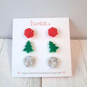 May include: A set of four pairs of stud earrings on a white card. The earrings include red flower shapes, green Christmas trees, and silver glitter circles. The card has the word "twee" in pink and the website address www.TweeAccessories.etsy.com.