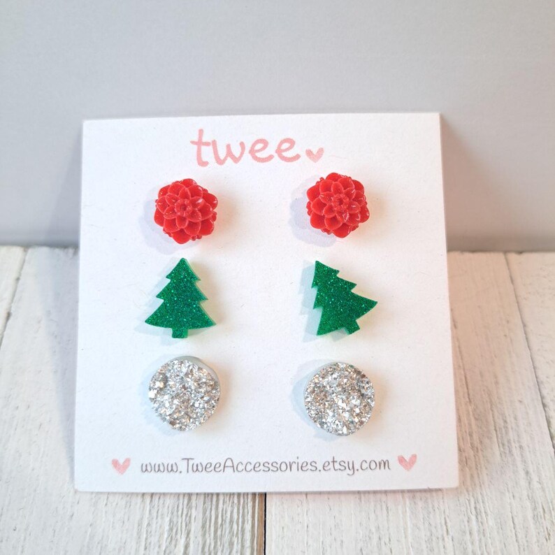 May include: A set of four pairs of stud earrings on a white card. The earrings include red flower shapes, green Christmas trees, and silver glitter circles. The card has the word "twee" in pink and the website address www.TweeAccessories.etsy.com.