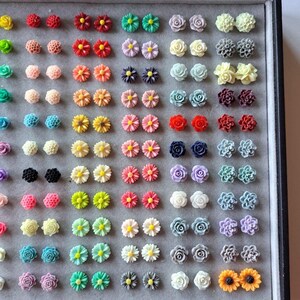 May include: A collection of colorful floral stud earrings in various designs, including roses, daisies, and other flower shapes. The earrings are arranged in a gray display case, showcasing a variety of colors such as red, yellow, pink, and blue.