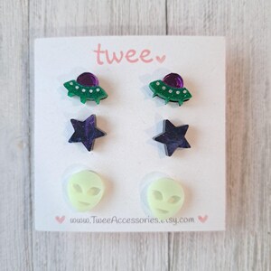 May include: A set of six stud earrings on a white card. The earrings feature three different designs: green UFOs with purple domes, dark purple stars, and pale green alien faces. The card has the word "twee" in pink script.