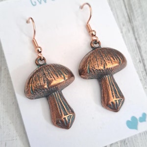 Close up of mushroom earrings in copper with a verdigris patina and bright copper ear wires. Shown on a white earring card that says Twee and has 2 little hearts in the lower right corner. Text and hearts are aqua colored.