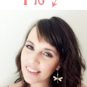 May include: A woman with long brown hair and a smile is wearing a pair of gold dragonfly earrings with a pearl accent.