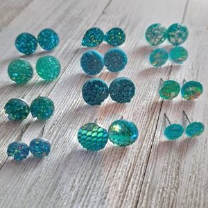 May include: A collection of teal and turquoise stud earrings in various designs. The earrings have textured, glittery, and iridescent surfaces. Some are round, others teardrop-shaped, and some have a scale-like pattern. The earrings are displayed on a light wood surface.