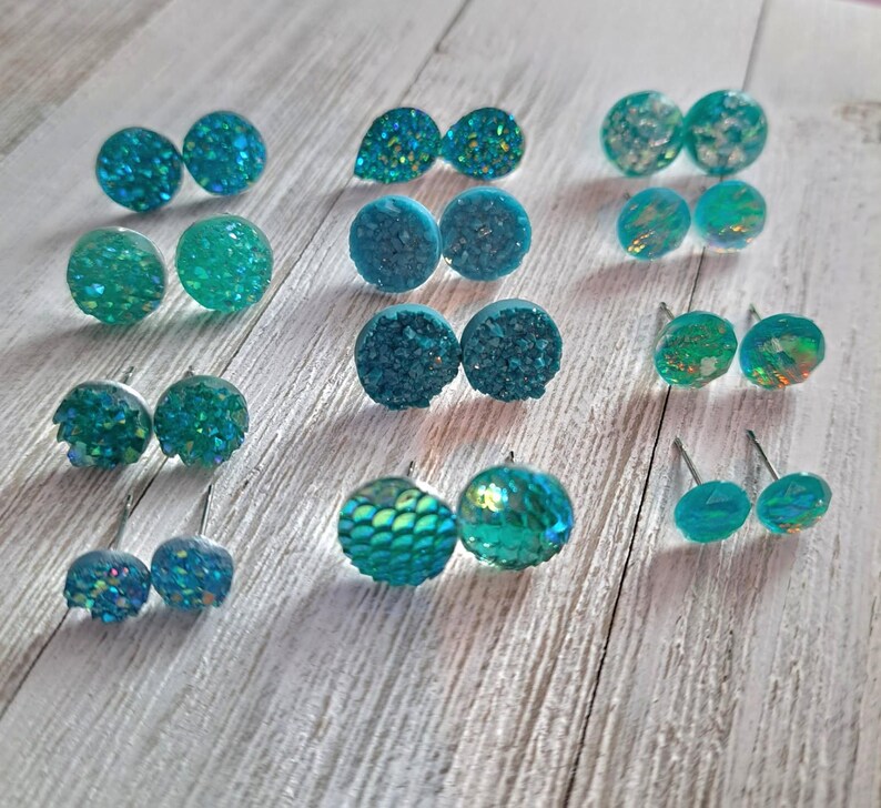 May include: A collection of teal and turquoise stud earrings in various designs. The earrings have textured, glittery, and iridescent surfaces. Some are round, others teardrop-shaped, and some have a scale-like pattern. The earrings are displayed on a light wood surface.