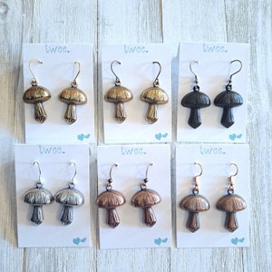 Six pairs of mushroom earrings each on a white earring card. Photo shows mushrooms in antiqued silver, antiqued brass with gold wires, antiqued brass with brass wires, antiqued copper, copper verdigiris, and matte black.