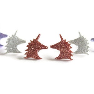 May include: Glittery unicorn head earrings in purple, silver, and red-orange. The earrings are arranged in a semi-circle, each with a horn and detailed mane. The earrings are small, likely stud earrings, and are set against a white background.